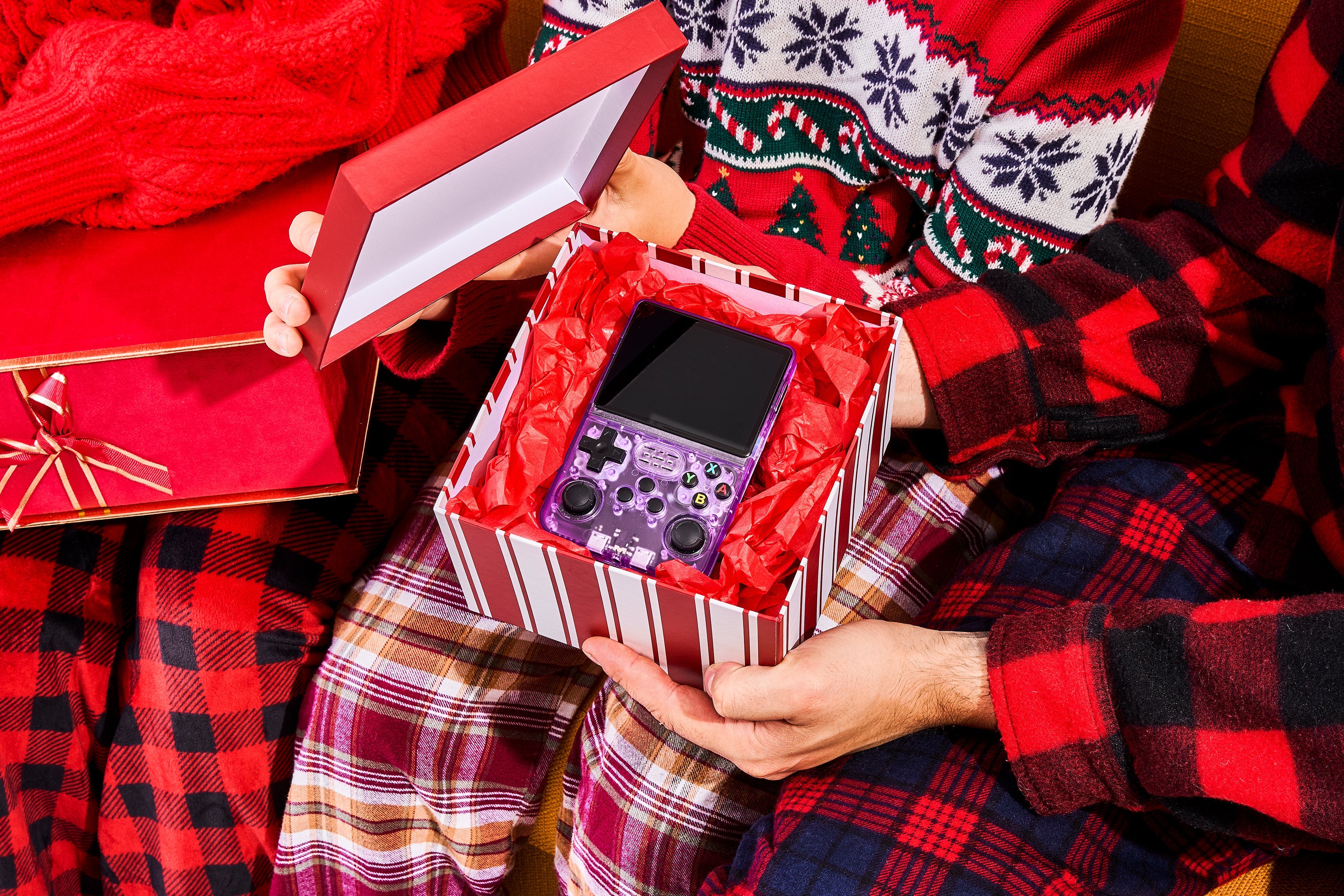 Person opening a gift box with a r36s retro handheld video game console inside, wearing festive pajamas.